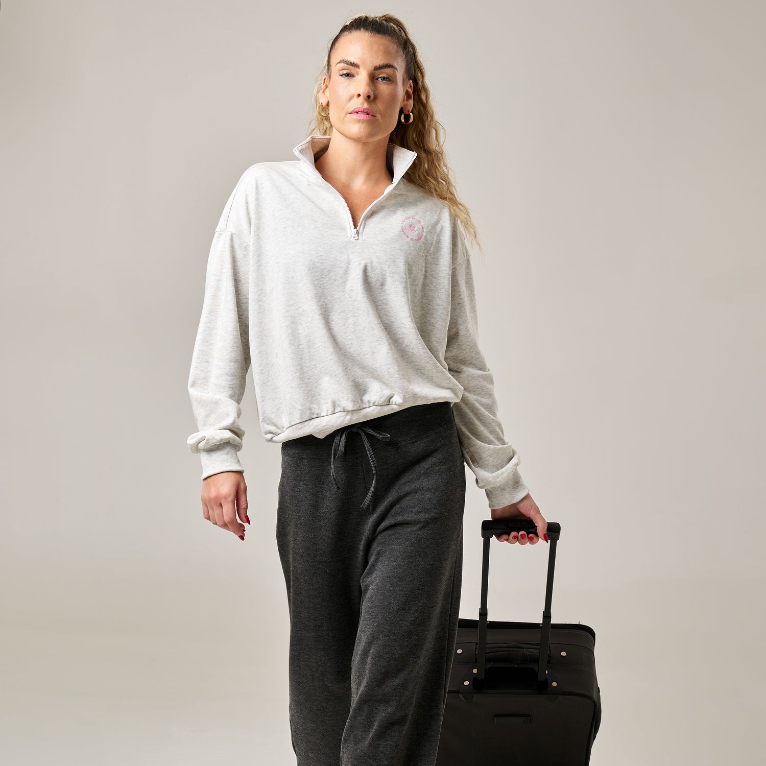 Woman in gray sweatshirt and dark pants holding a black suitcase against a plain background